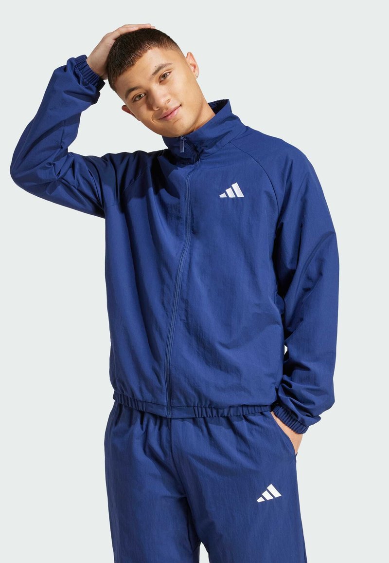 Marineblå zip-up jacka i lätt material, med elastiska ärmslut och midja, samt en vit logotyp på bröstet.