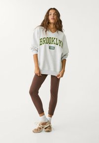 Grå oversized sweatshirt med "BROOKLYN 1986" i grönt, tillsammans med bruna leggings och neutralt färgade sneakers. Texturerad tyg.