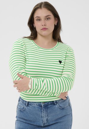 Kvinde med langt brunt hår iført grøn og hvidstribet langærmet skjorte med lille sort hjerte, blå jeans og sølvsmykker, med armene over kors.