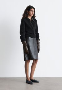 Schwarze, flauschige Strickjacke mit rundem Kragen, Knopfleiste und strukturierten Ärmeln, kombiniert mit einem knielangen Rock aus schwarzem Krokodilprägeleder. Braune Handschuhe.