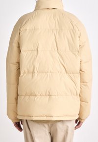 Chaqueta de plumas beige con cuello alto, textura acolchada y puños elásticos. La vista trasera muestra costuras horizontales y un ajuste holgado.