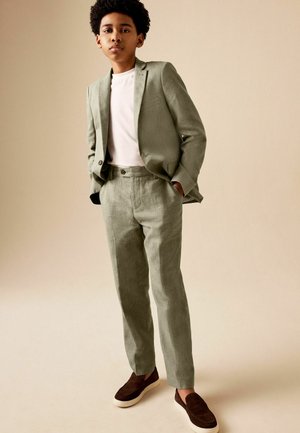 Junge trägt eine hellgraue Anzugjacke und Hose mit einem weißen Hemd und dunkelbraunen Slippern, steht mit den Händen in den Taschen.