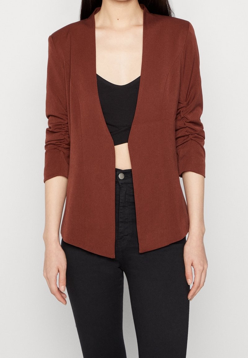 Hnědý blazer s otevřeným předním zapínáním, dlouhými rukávy a shromážděnými detaily. Nositý přes černý crop top, spárovaný s tmavými džínami, s minimálním designem.