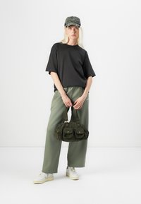 Schwarzes übergroßes T-Shirt, hellgrüne weite Hosen, olivgrüne Cordtasche mit Taschen und weiße Sneakers. Mütze mit strukturierter Oberfläche.