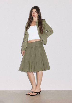 Femme portant une veste et une jupe vert olive avec un crop top blanc et des sandales noires à talons, debout sur un fond gris clair.