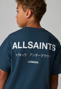 Jūro mėlynas medvilninis marškinėliai su ryškiai balta užrašais: "ALLSAINTS" ir "LONDONYJE" bei žemiau esančiais japonų simboliais. Trumpomis rankovėmis ir apvaliu iškirpimu.