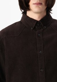 Camicia di cotone a coste marrone con bottoni sul davanti, colletto a punta e righe verticali testurizzate. Presenta bottoni neri lungo la patta.