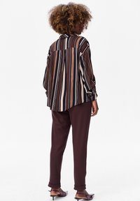 Vrouw met krullend haar die een losse blouse met verticale strepen in bruin, zwart en beige draagt, donderbruine broek en paarse schoenen met hak, met de rug naar voren.