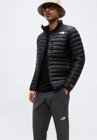 Άνδρας που φοράει μπεζ καπέλο τύπου bucket, γυαλιά ηλίου, μαύρο φουσκωτό μπουφάν και σκούρο γκρι παντελόνι, στέκεται με το ένα χέρι στην τσέπη μπροστά σε απλό φόντο.