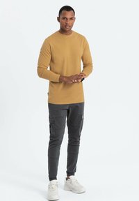 Langärmliges senfgelbes Hemd kombiniert mit dunklen Cargo-Hosen und weißen Sneakers. Das Hemd hat einen Rundhalsausschnitt; die Hose verfügt über mehrere Taschen.