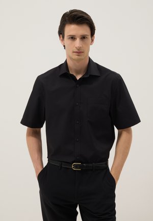Junger Mann mit dunklem Haar, der ein schwarzes kurzärmeliges Hemd mit Knöpfen und schwarze Hose trägt, steht mit den Händen in den Taschen vor einem schlichten Hintergrund.