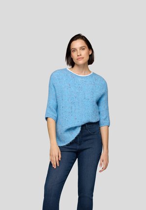 Frau trägt einen hellblauen, gesprenkelten Strickpullover mit hochgerollten Ärmeln und dunklen blauen Jeans, stehend vor einem schlichten hellgrauen Hintergrund.