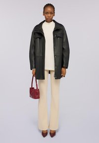 Chaqueta de cuero negra con dos bolsillos frontales, blusa beige y pantalones de campana color crema. Sostiene un pequeño bolso rojo y lleva zapatos de tacón marrones.