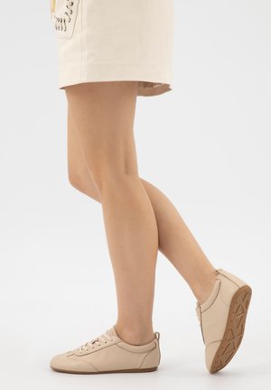 Benenden die beige lage sneakers met gumzolen dragen, gecombineerd met een lichtbeige minirok met zijveters, op een witte achtergrond.