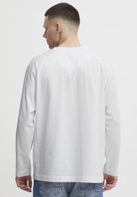 Weißes Langarmhemd mit glatter Textur, Raglanärmeln und einem kleinen geprägten Logo auf der Rückseite, nahe dem Kragen. Getragen mit blauen Jeans.
