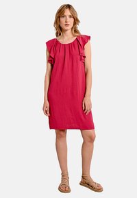 Abito corto fucsia con maniche a cappuccio arricciate, vestibilità ampia, lunghezza al ginocchio e texture morbida. Indossato con sandali piatti con cinturini.