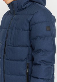 Marineblaue Steppjacke mit gestepptem Design, hoher Kragen, Reißverschlusstaschen und einem auffälligen Logo-Patch am Ärmel.