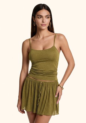Donna che indossa un top verde oliva senza maniche e una gonna abbinata con orecchini a cerchio dorati e braccialetto, in piedi davanti a uno sfondo semplice.