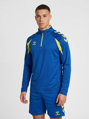 Ung mand iført en blå og gul sportstop med kvart lynlås og matchende shorts, stående mod en ensfarvet lys baggrund.