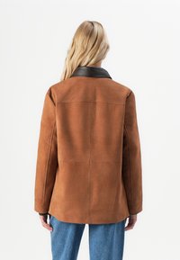 Persona con lunghi capelli biondi che indossa una giacca scamosciata marrone con colletto in pelle nera e jeans blu, mostrata di spalle.
