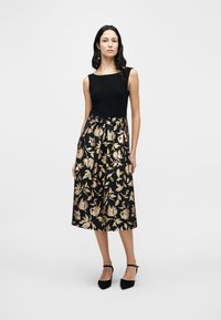 Μαύρο αμάνικο τοπ συνδυασμένο με φούστα μήκους μέχρι το γόνατο, διακοσμημένη με χρυσό λουλουδάτο μοτίβο σε μαύρο φόντο. Το μοντέλο φοράει μαύρα τακούνια.