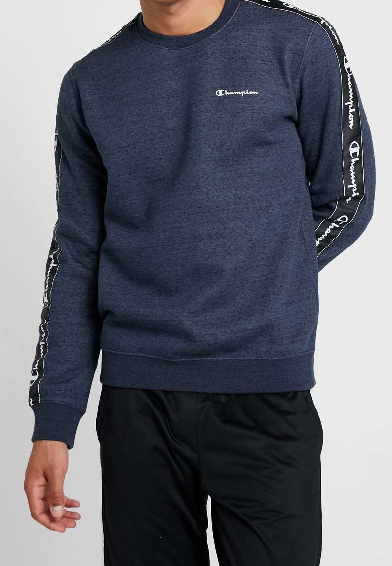Homme portant un sweat-shirt Champion bleu marine avec logo sur la poitrine et bande de marque le long des manches, associé à un pantalon noir.