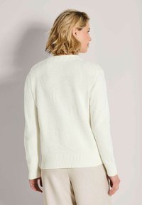 Vrouw met schouderlang blond haar, gekleed in een witte gebreide trui en lichtbeige broek, met de rug naar de camera tegen een effen achtergrond.