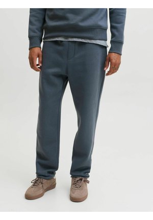 Personne portant un pantalon de survêtement gris foncé, des chaussures décontractées marron, et un sweat-shirt gris foncé assorti, debout sur un fond uni.