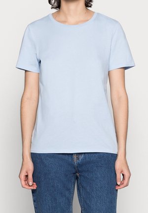 Person iført en ensfarvet lyseblå kortærmet T-shirt og blå denimjeans, stående mod en neutral baggrund.