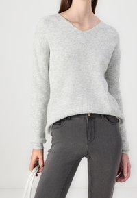 Suéter gris de punto acanalado con escote en V y mangas largas, combinado con jeans gris oscuro. La modelo sostiene un bolso blanco, mostrando un atuendo casual.