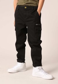 Zwarte cargobroek met zijritsen, een snug fit, geribbelde zoom en een gele knoopaccent. Gecombineerd met witte high-top sneakers.