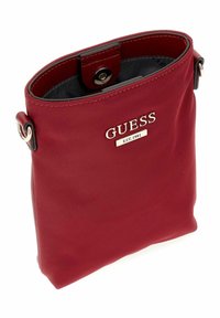 Sac à main en nylon rouge à texture lisse, doté d'une fermeture à pression et d'accents argentés. Le nom de la marque "GUESS" est affiché en évidence.