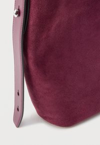 Vínovočervená semišová kabelka s hladkým koženým lemovaním, zaobleným tvarom a strieborným detailom so zapínaním na bočnej strane. Viditeľný textúrovaný povrch.