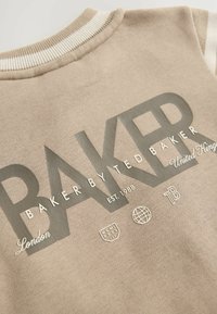 Bežsviiter ribidega kaelusega, millel on julge hall tekst "BAKER BY TED BAKER" ja erinevad väikesed embleemid ees.