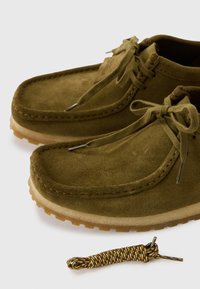 Zapatos de ante verde olivo con punta redondeada y cordones a juego. Suela de goma con un agarre texturizado. Incluye un cordón de repuesto con un diseño.