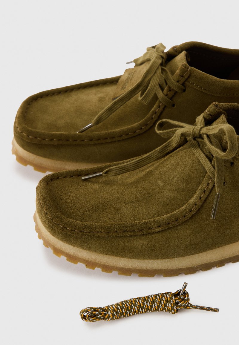 Zapatos de ante verde olivo con punta redondeada y cordones a juego. Suela de goma con un agarre texturizado. Incluye un cordón de repuesto con un diseño.