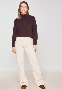 Pull en tricot bordeaux avec un col montant et des accents en boutons, associé à un pantalon évasé crème. Des chaussures marron complètent la tenue.