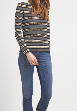 Frau trägt ein schwarz-weißes Langarmshirt mit Goldketten- und Punktemustern, kombiniert mit enganliegenden blauen Jeans.