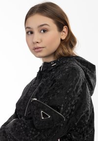 Giacca nera con cappuccio che presenta un motivo geometrico testurizzato, una chiusura zip frontale e una tasca con zip triangolare sulla manica. Materiale leggero.