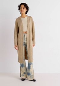 Beige stickad cardigan med öppen front och långa ärmar, tillsammans med batikmönstrade vida byxor i blått och gräddvitt. Svarta skor.