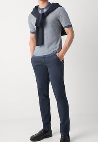 Chino blu scuro abbinati a una polo a righe a maniche corte. Un maglione scuro è appoggiato sulle spalle. Mocassini neri completano il look.