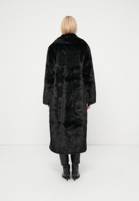 Svart kåpeliknende jakke i kunstig pels med lang silhuett, høy krage og brede ermer, satt mot en nøytral bakgrunn. Glatt tekstur og luksuriøst utseende.