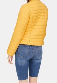 Giacca trapuntata gialla con collo alto e maniche lunghe, abbinata a pantaloni pantaloncini in denim blu. La giacca presenta cuciture orizzontali e un design aderente.
