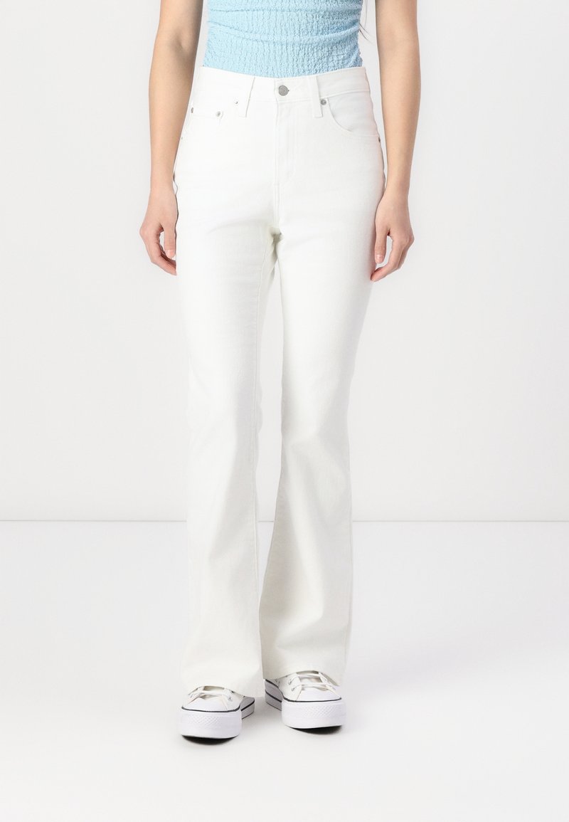 Personne portant un jean blanc flare taille haute, un top sans manches bleu clair texturé et des baskets blanches à plateforme sur fond blanc uni.