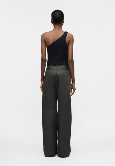 Γυναίκα με την πλάτη να φαίνεται, φορώντας μαύρο τοπ με έναν ώμο και χαλαρό ανοιχτό γκρι παντελόνι με φαρδιά μπατζάκια, μπροστά σε απλό φωτεινό φόντο.