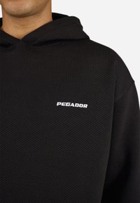Sort tekstureret hoodie med hvid "PEGADOR" logo broderet på venstre bryst, båret af en person vist fra hals til skulder.
