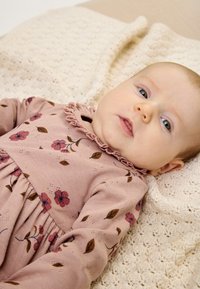 Hosszú ujjú, világos rózsaszín ruha virágmintával és fodros nyakkivágással. Texturált krém színű takarón, kötött mintával van elhelyezve.