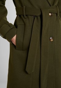 Olijfgroene jas van gestructureerde stof, met een ceintuur in de taille, een knoopsluiting en zijzakken.