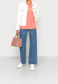 Kobieta w białej dżinsowej kurtce, koralowej koszulce z grafiką, niebieskich szerokich dżinsach, białych trampkach, trzymająca u swojego boku różową torebkę.