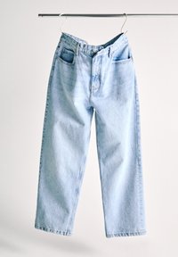 Jean en denim bleu clair suspendu sur un cintre blanc contre un fond blanc uni, avec le bouton supérieur défait.